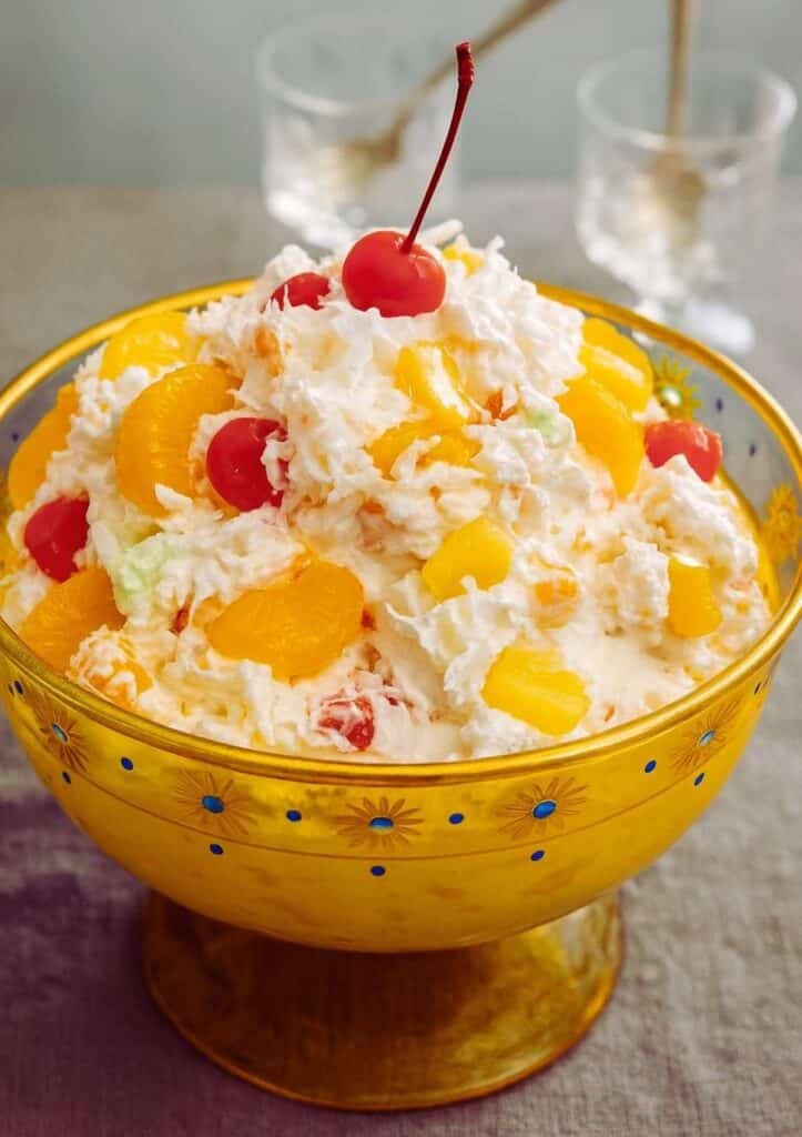classic ambrosia salad in yellow glass bowl topped with cherries, pineapple, and coconut