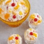 top view of creamy ambrosia salad with cherries, pineapple, and oranges in a yellow glass bowl and small dessert cups