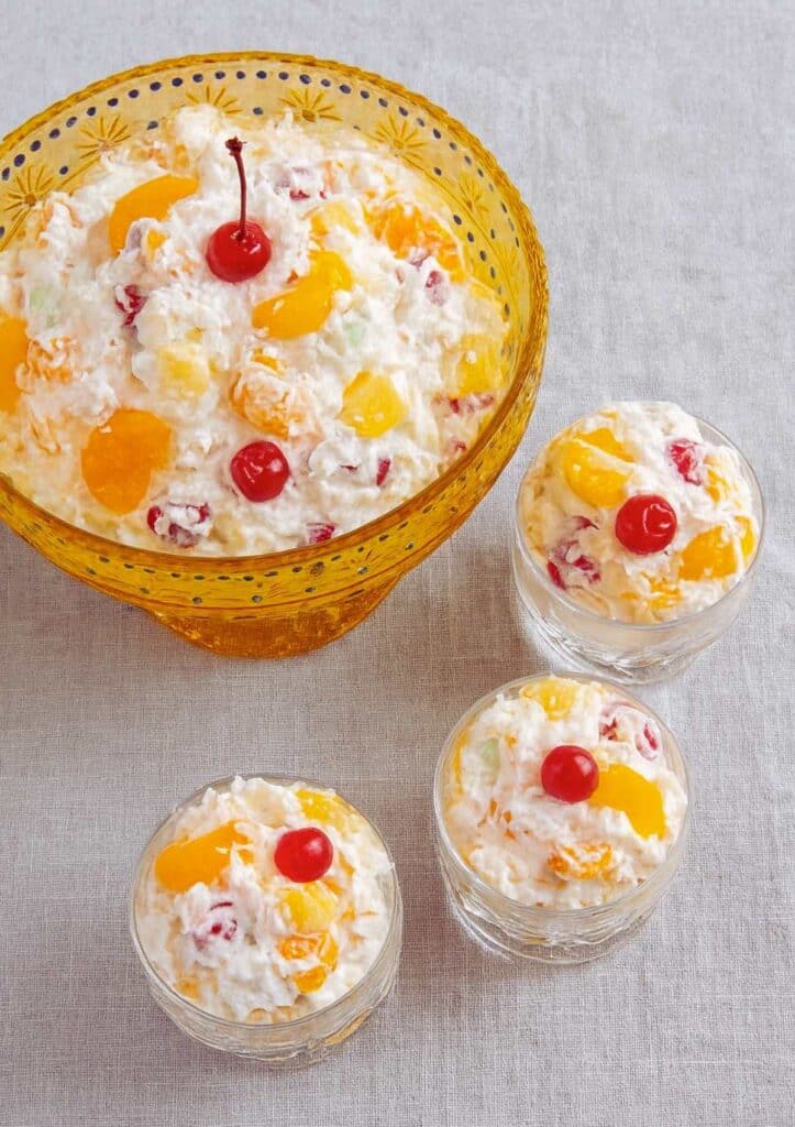 top view of creamy ambrosia salad with cherries, pineapple, and oranges in a yellow glass bowl and small dessert cups