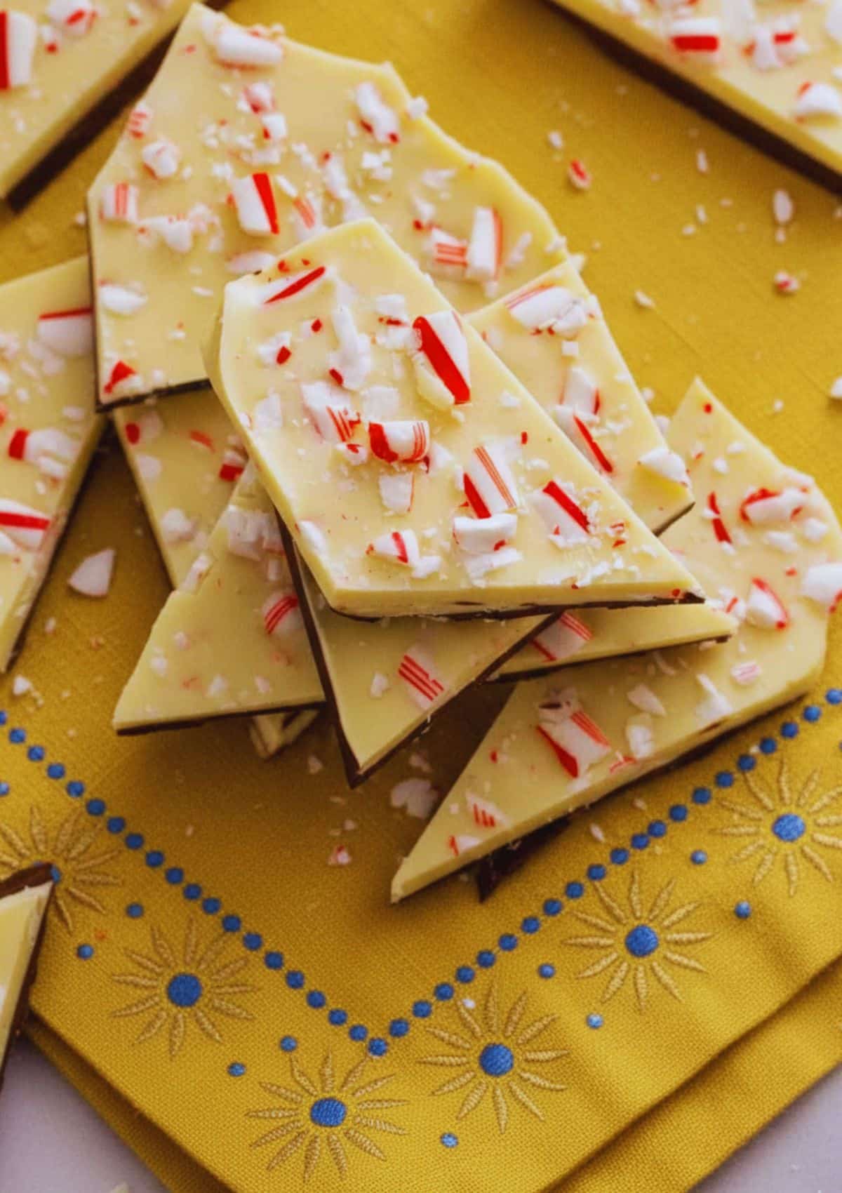 Peppermint bark pieces with crushed candy canes stacked on a yellow patterned cloth
